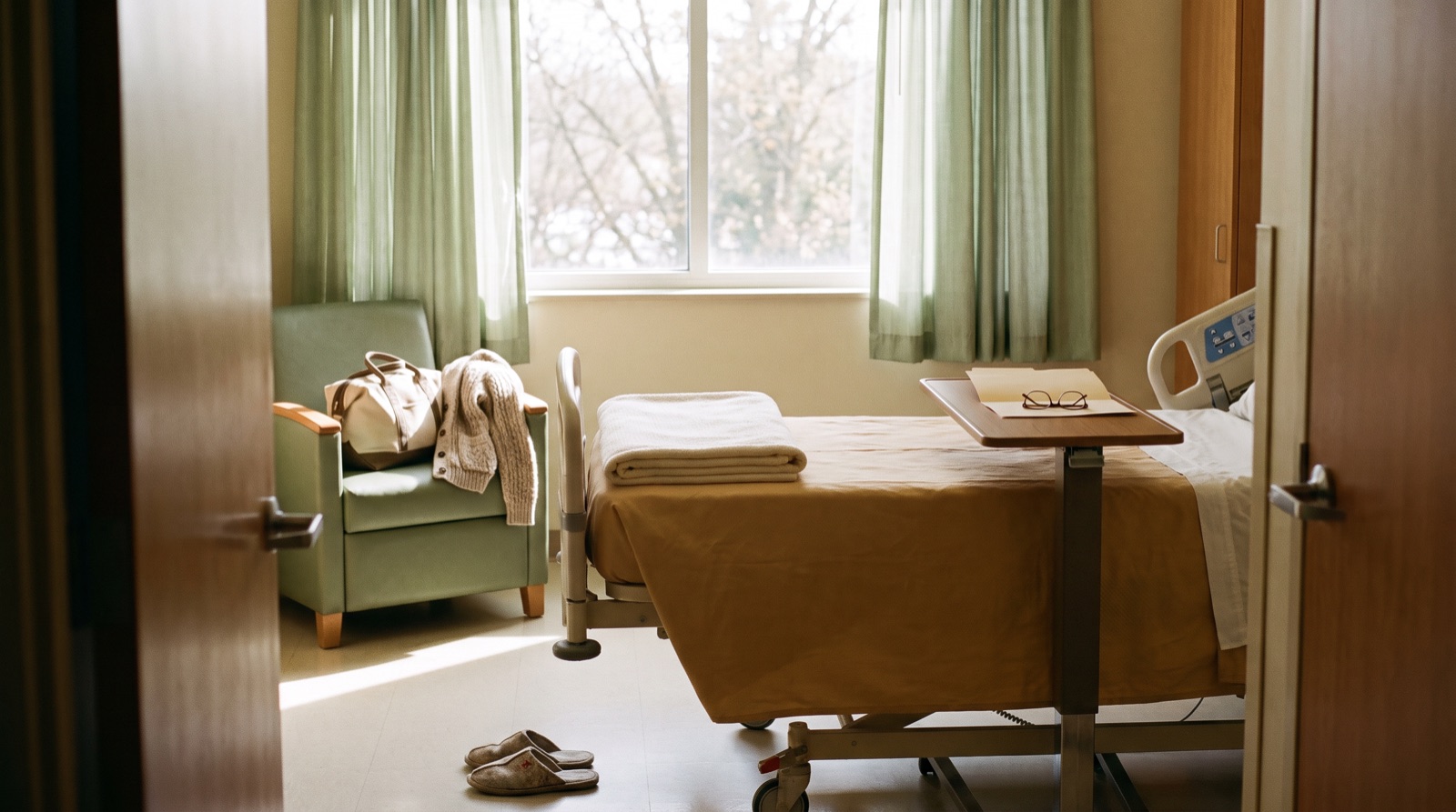 An empty, sunlit hospital room on discharge day with a folded blanket on the bed, a packed bag on the visitor chair and a closed folder on the bedside table.