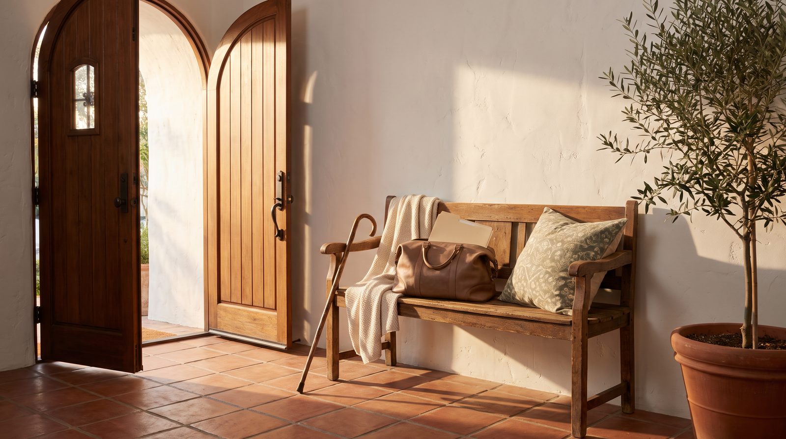 A sunlit Spanish-style entry hall with a bench holding a travel bag, a folder, a folded blanket and a pillow, a cane resting beside it and the front door open.