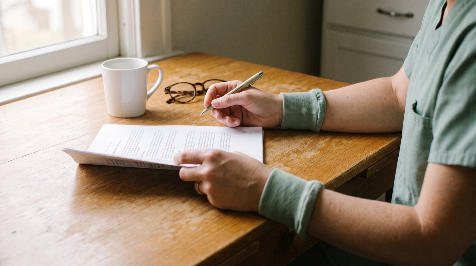A nurse in sage green scrubs sitting at a wooden kitchen table with a pen, reviewing a printed care plan beside a mug and a pair of glasses, lit by a window.