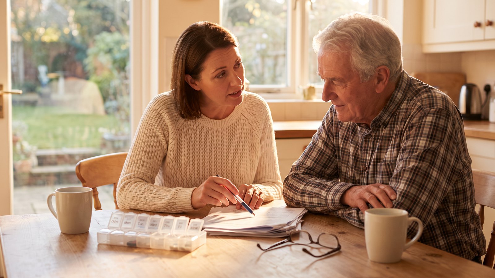 An adult daughter and her father sit together at a kitchen table reviewing hospital discharge paperwork in warm afternoon light.